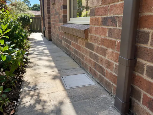 Paved side path beside a brick house with a square drain cover and dry walkway in Turramurra